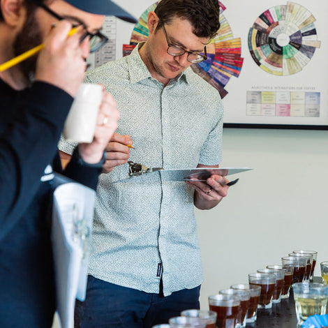 Two men evaluating a row of cups of coffee. One is sipping from a spoon and one is taking notes on a clipboard.