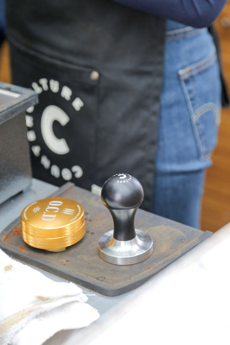 Photo of an espresso tamper and other tools on a table. A person wearing a Counter Culture Coffee apron is in the background. 