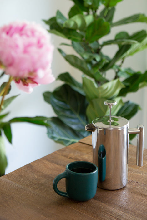 Photo of a mug and French press on a wooden surface with a peony in the foreground. 
