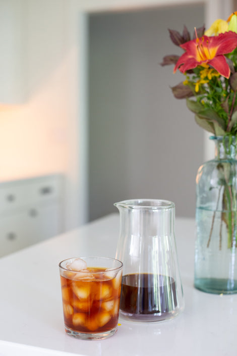 Photo of a carafe of cold coffee next to a glass of iced coffee on a kitchen counter. 